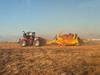 A red tractor pulls a yellow earthmover across a dry field under a clear blue sky.
