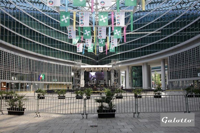 Courtyard with stage, banners, and modern glass building. Metal barriers and potted plants in foreground.