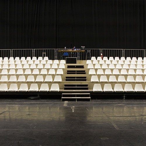 Rows of empty white chairs face a raised stage with a desk and black backdrop.