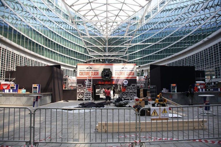 Event setup in a modern building with glass roof; barriers, stage, equipment, and workers are visible.