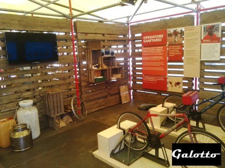 Interior with wooden pallet walls, TV, bicycle-powered generator, educational posters, and containers, possibly an exhibit.