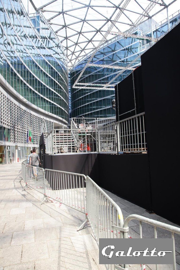Exterior view of a modern building with a performance stage set up outside. Metal barriers and a black backdrop.