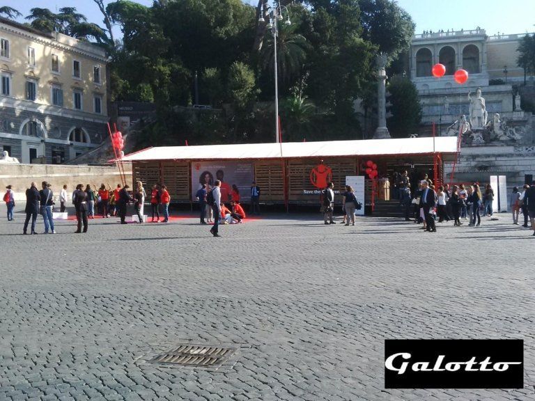 People in a plaza with a temporary structure, possibly an event space. Building and red balloons are in the background.