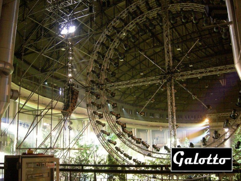 Large venue with a circular lighting rig, spotlights, and steel trusses, partially outdoors.