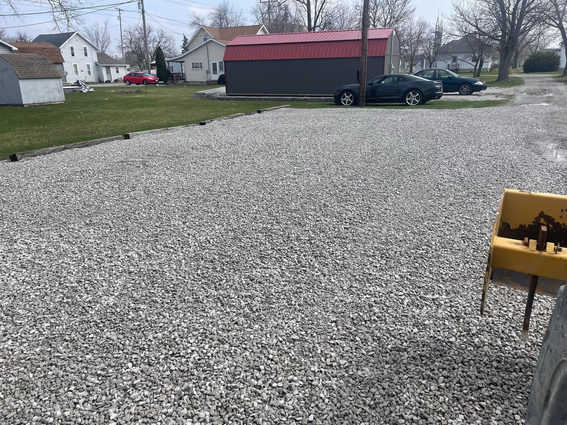 Gravel driveway with parked cars and buildings in the background. Green grass and a yellow object are also visible.