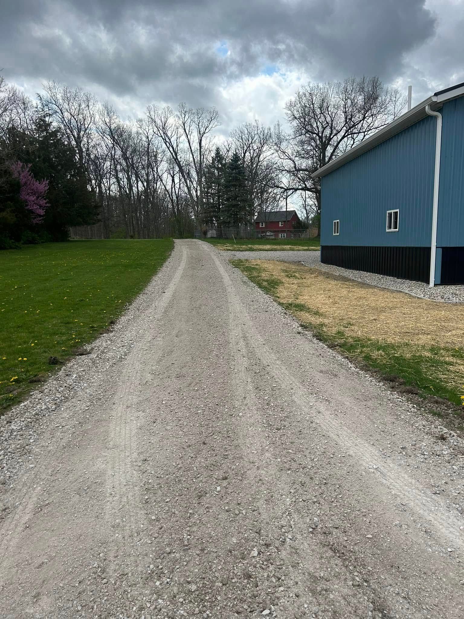 Gravel driveway leading toward bare trees and a blue building under a cloudy sky.