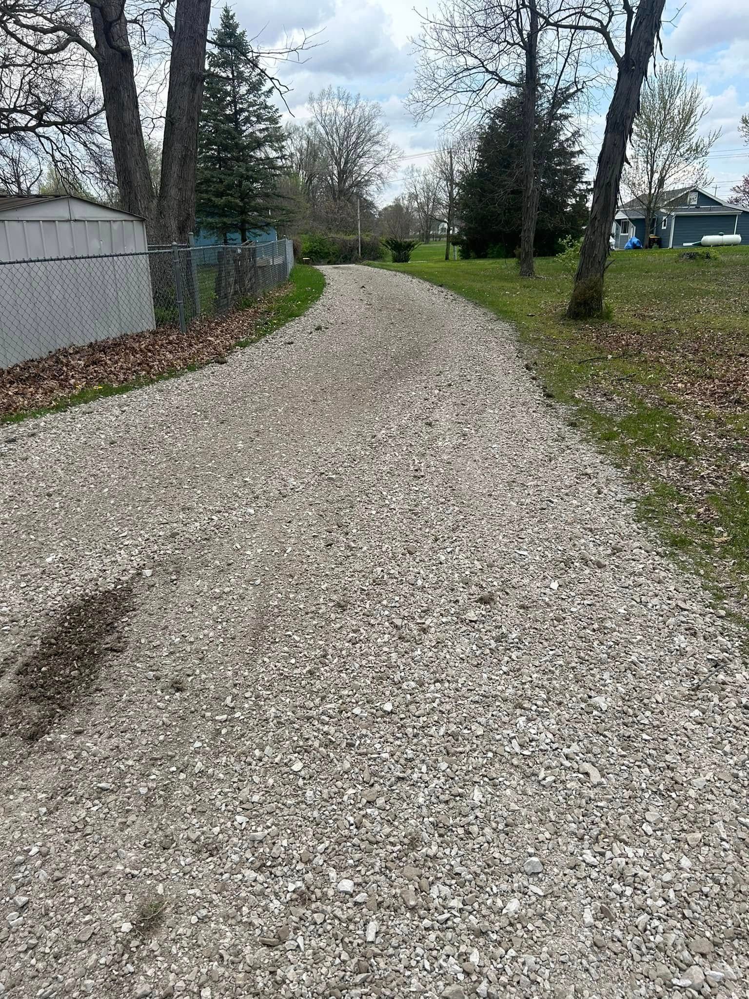 Gravel pathway leading toward trees, green grass, and houses under a partly cloudy sky.