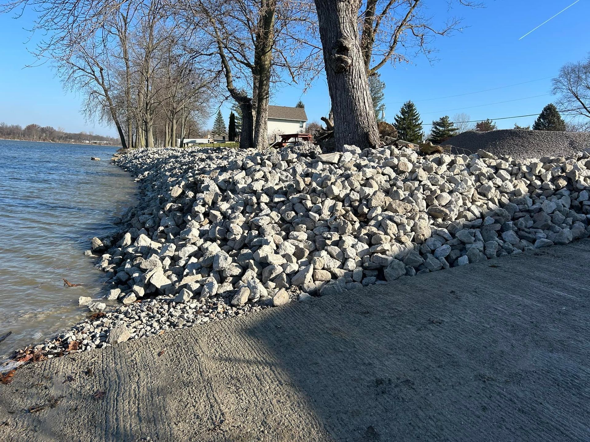 A shoreline reinforced with large rocks to prevent erosion. Trees and a house are in the background.