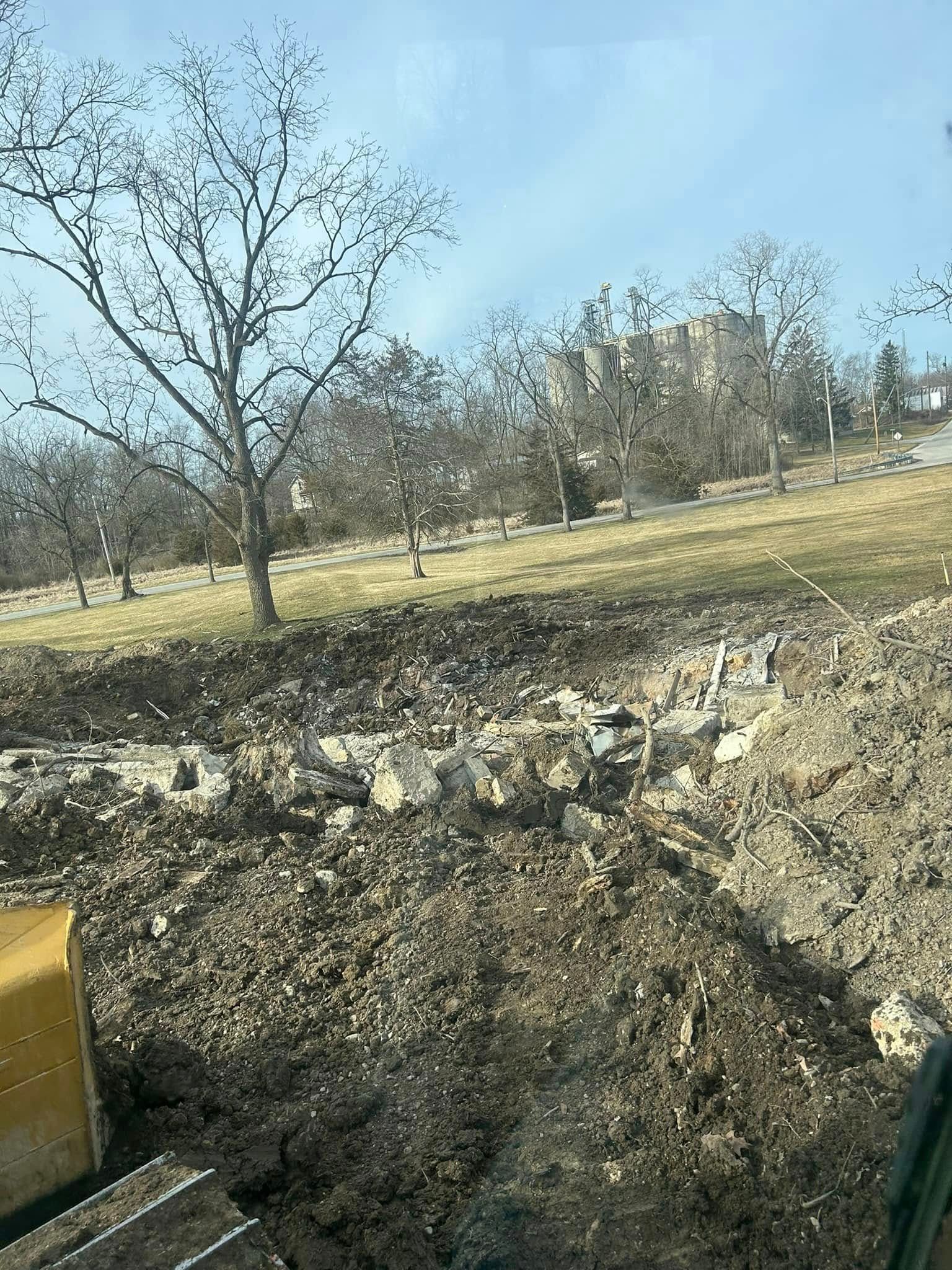 A muddy construction site with bare trees, grassy hill, and a building under a blue sky.