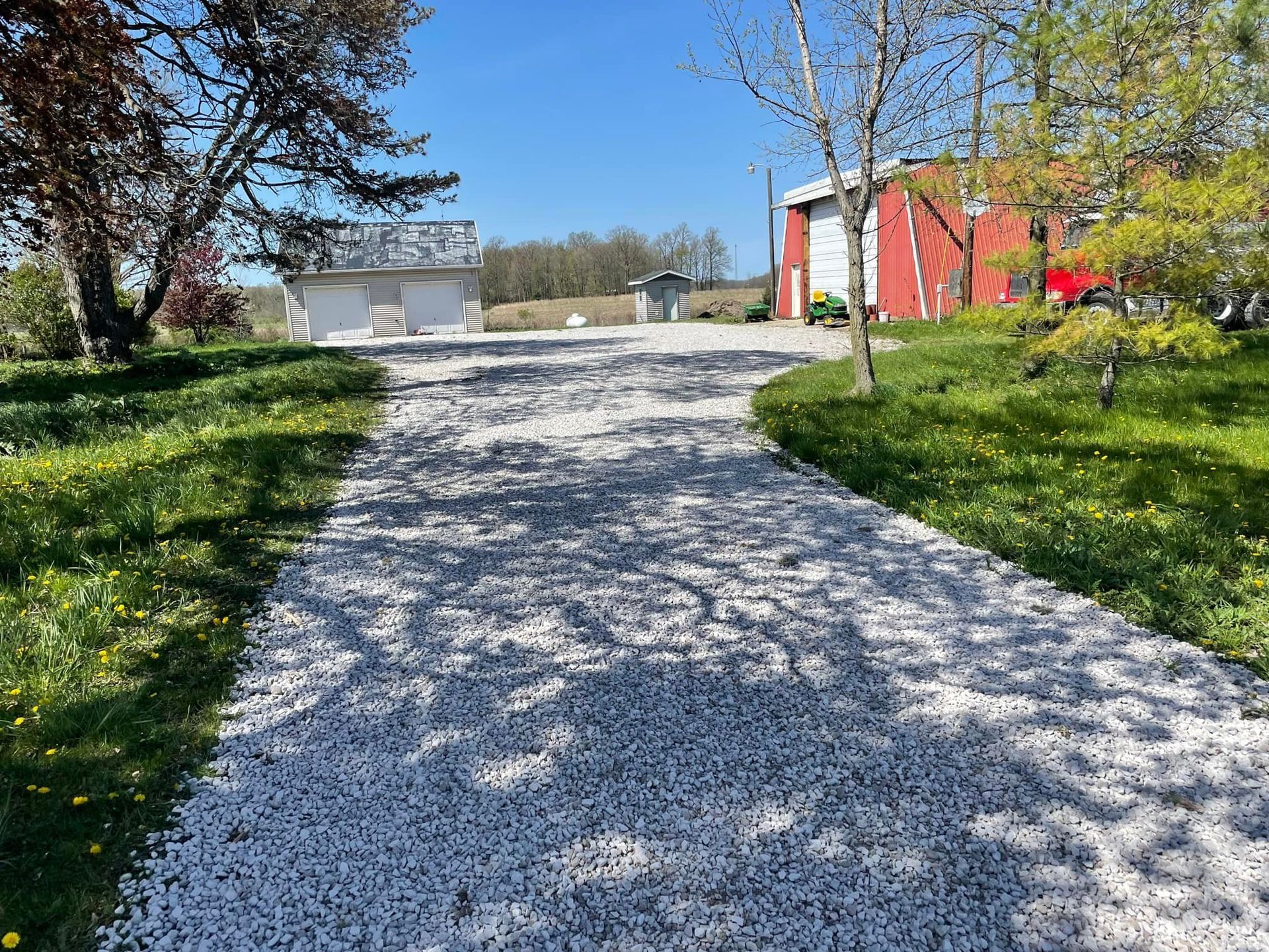 A gravel driveway leads to a white garage and red building under a blue sky, flanked by green grass and trees.