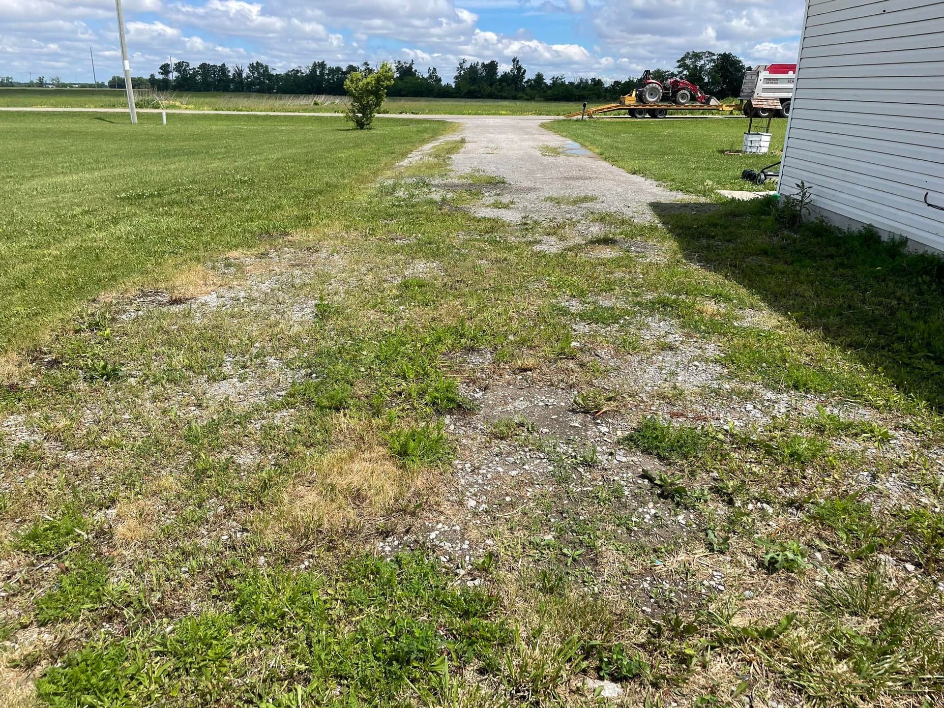 Gravel driveway leading to a grassy field. A white building is on the right side. Blue sky with trees in the background.