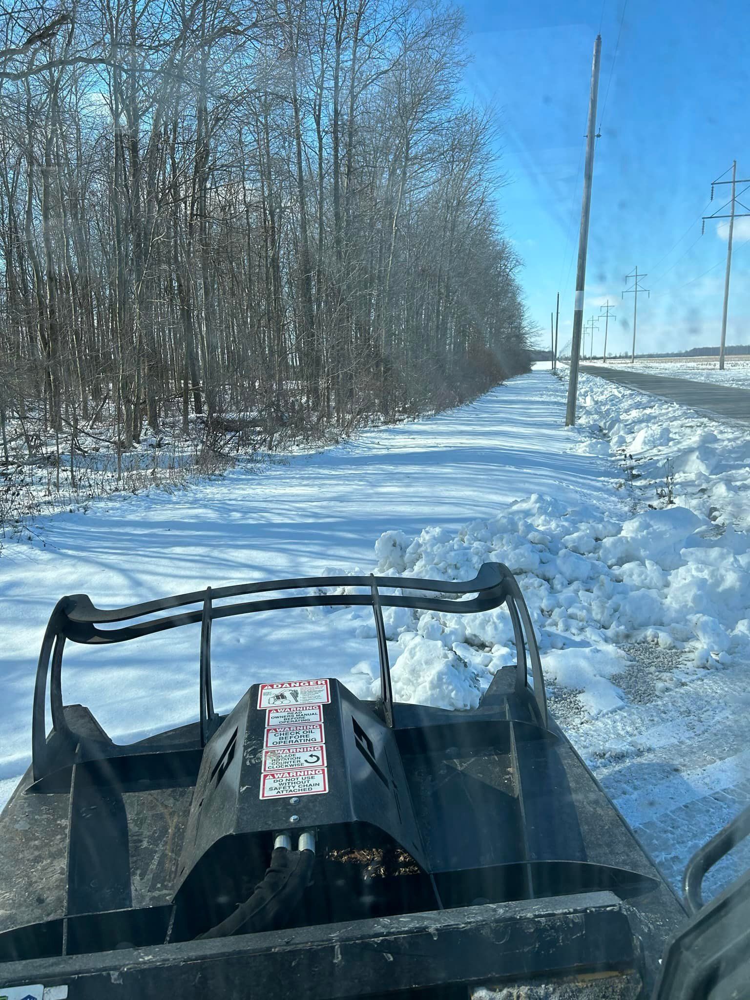 Skid steer clearing snow on a road next to a tree line and power poles.