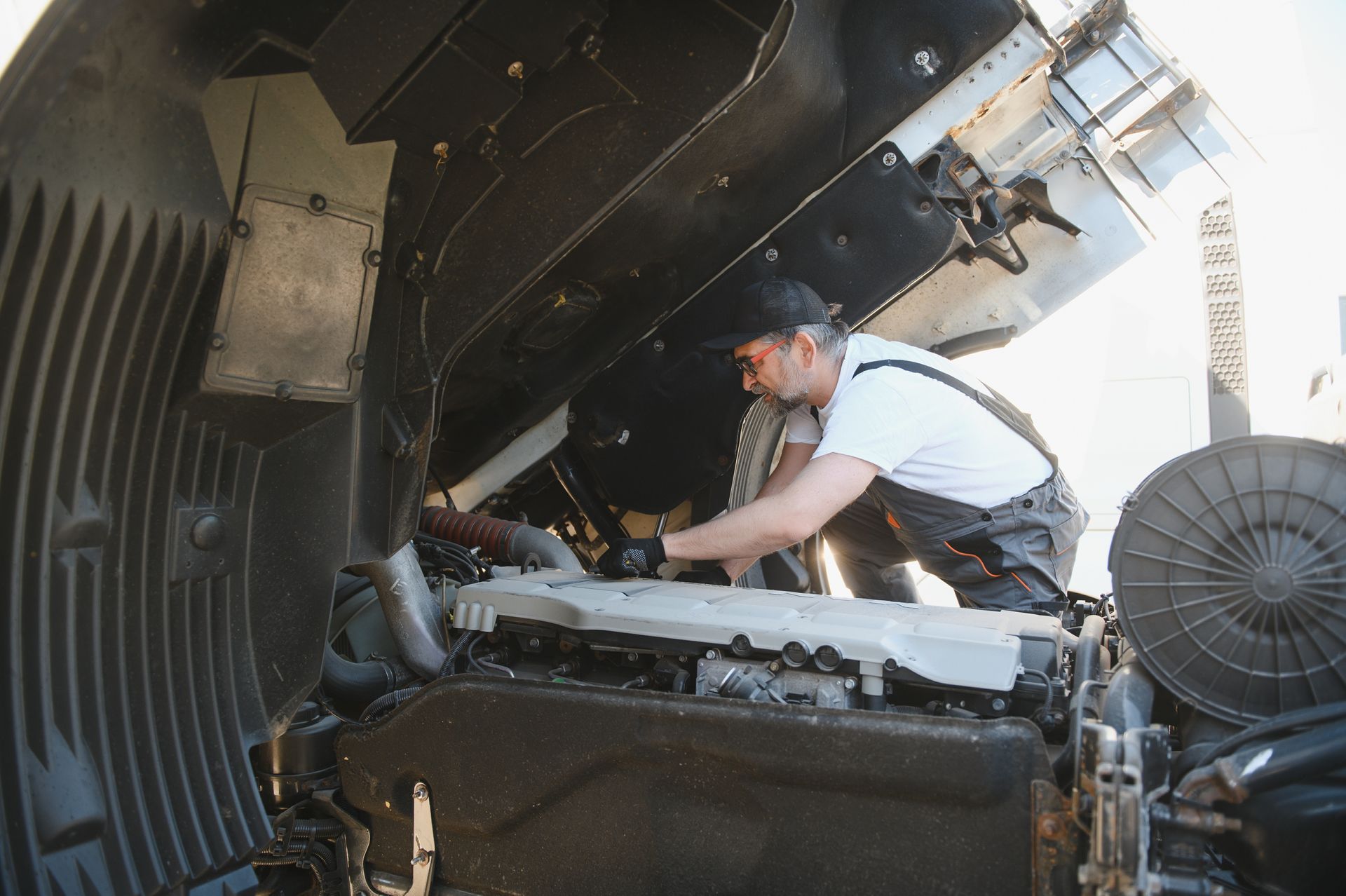Mechanic repairs truck engine with open hood. Outdoors, in daylight. Wearing work clothes and a cap.
