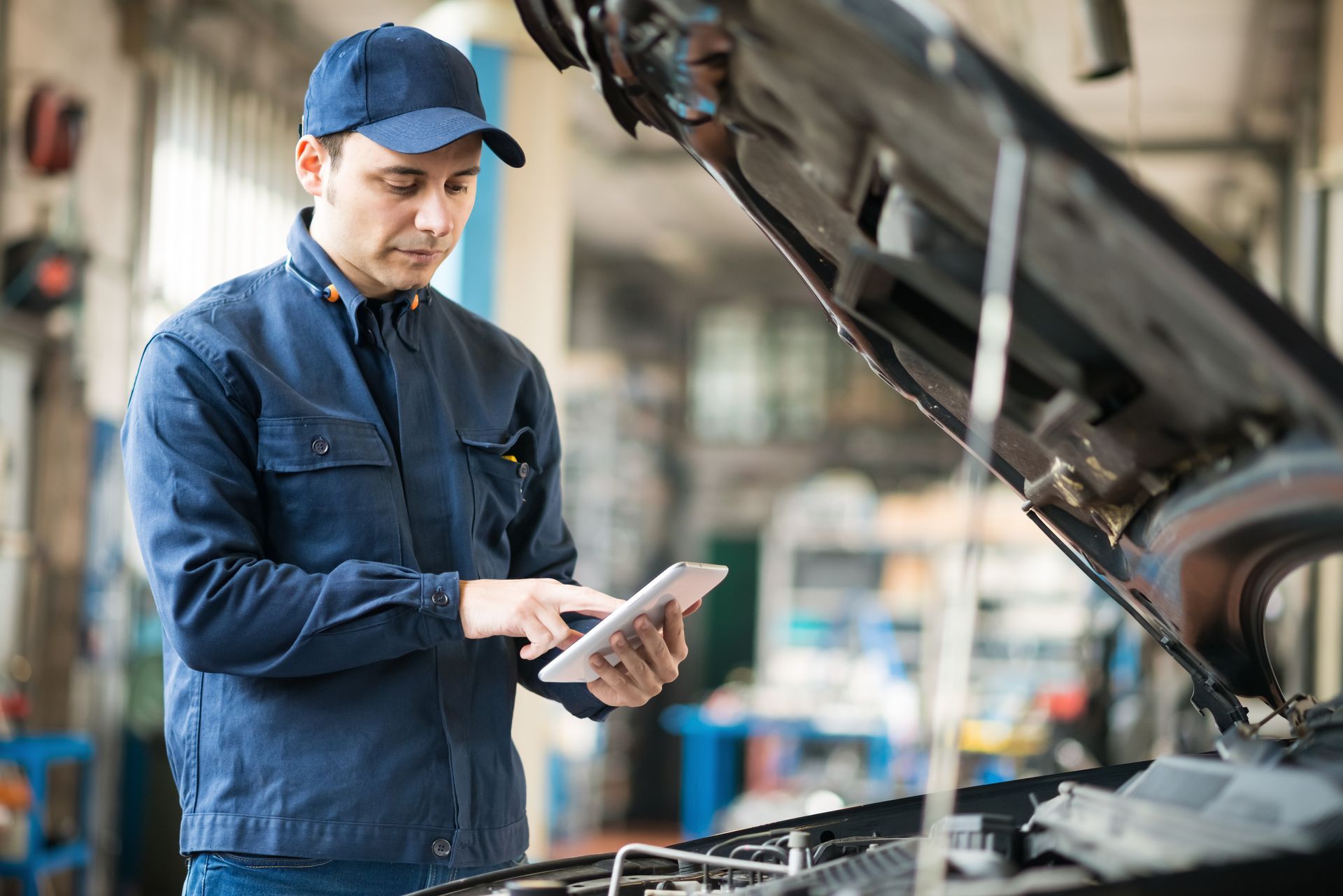 Mechanic in blue uniform using a tablet, looking at the car's open hood in a garage.