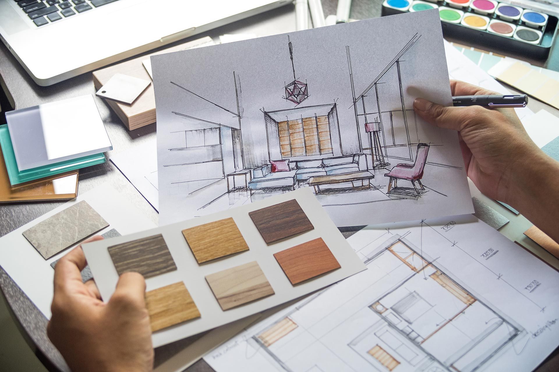 Hands holding an interior design sketch with material samples and floor plans on a desk