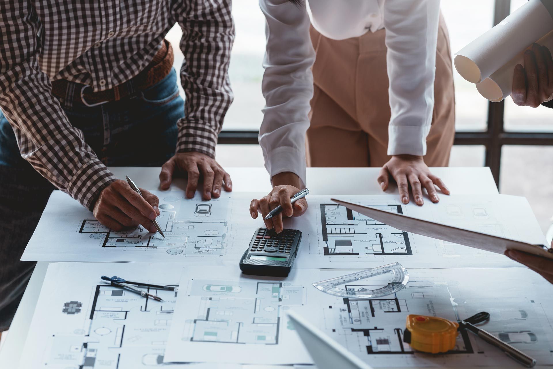 Two people review architectural floor plans and calculate costs at a desk with a calculator and measuring tools