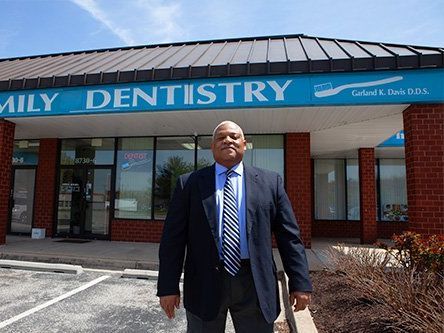 A man in a suit stands in front of "Family Dentistry" with a blue sign, brick building exterior, and a parking lot.