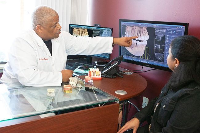 A dentist points at a dental x-ray on a monitor, explaining it to a seated patient. Both are in an office setting.