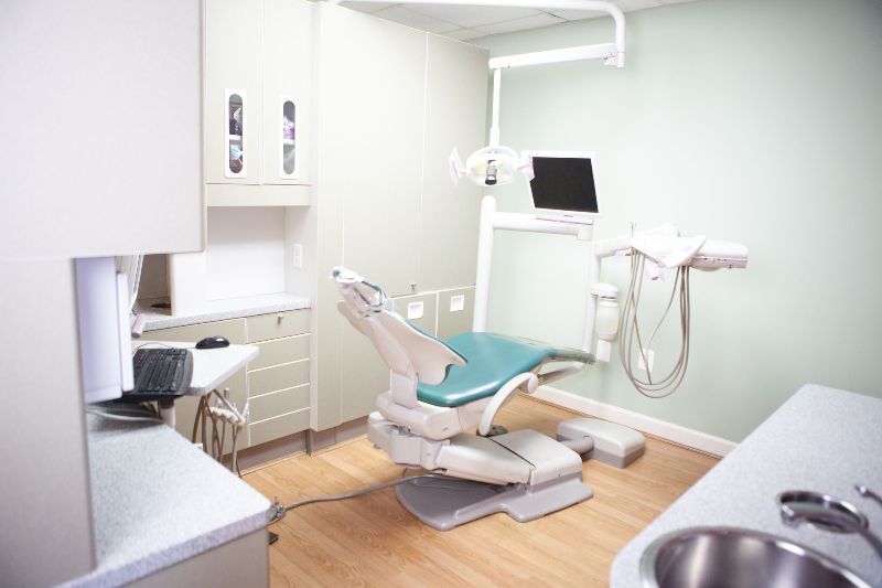 A dental office featuring a dental chair, equipment, cabinets, and a sink. The walls are light green, and the floors are wood-toned.
