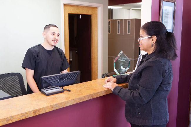 A male receptionist in black scrubs smiles and interacts with a woman at a front desk in a medical office.