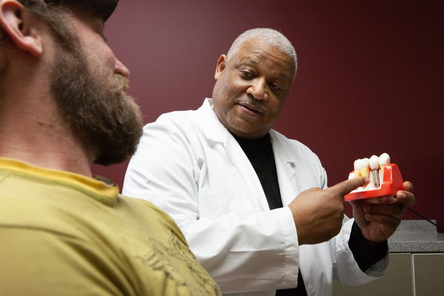 A Black doctor in a white coat points at a model of teeth, explaining to a patient with a beard. Interior, red background.