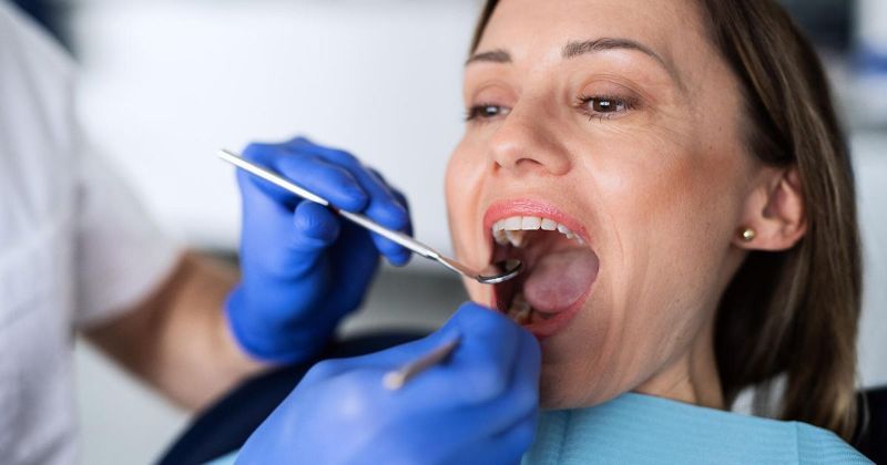 A dentist examines a woman's mouth with dental tools in a clinic. The woman opens her mouth, blue-gloved hands are visible.