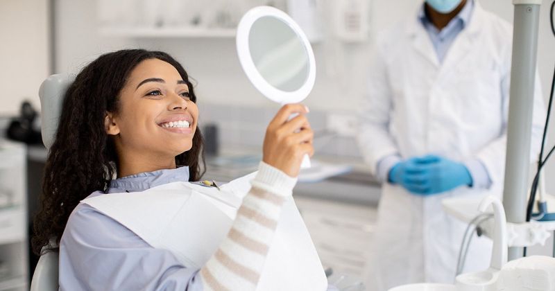 Woman smiles, admiring her teeth in a hand mirror at a dentist's office. A masked dentist stands in the background.