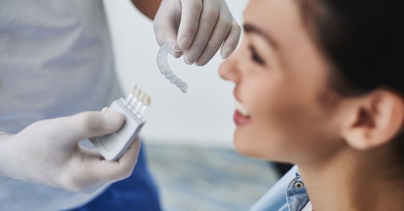A dentist, wearing gloves, holds a clear aligner near a smiling patient. Packages of aligners rest in his other hand.