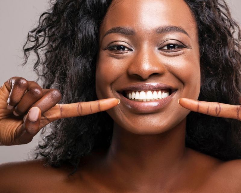 Woman with dark skin smiling widely, pointing to her teeth, against a neutral backdrop.