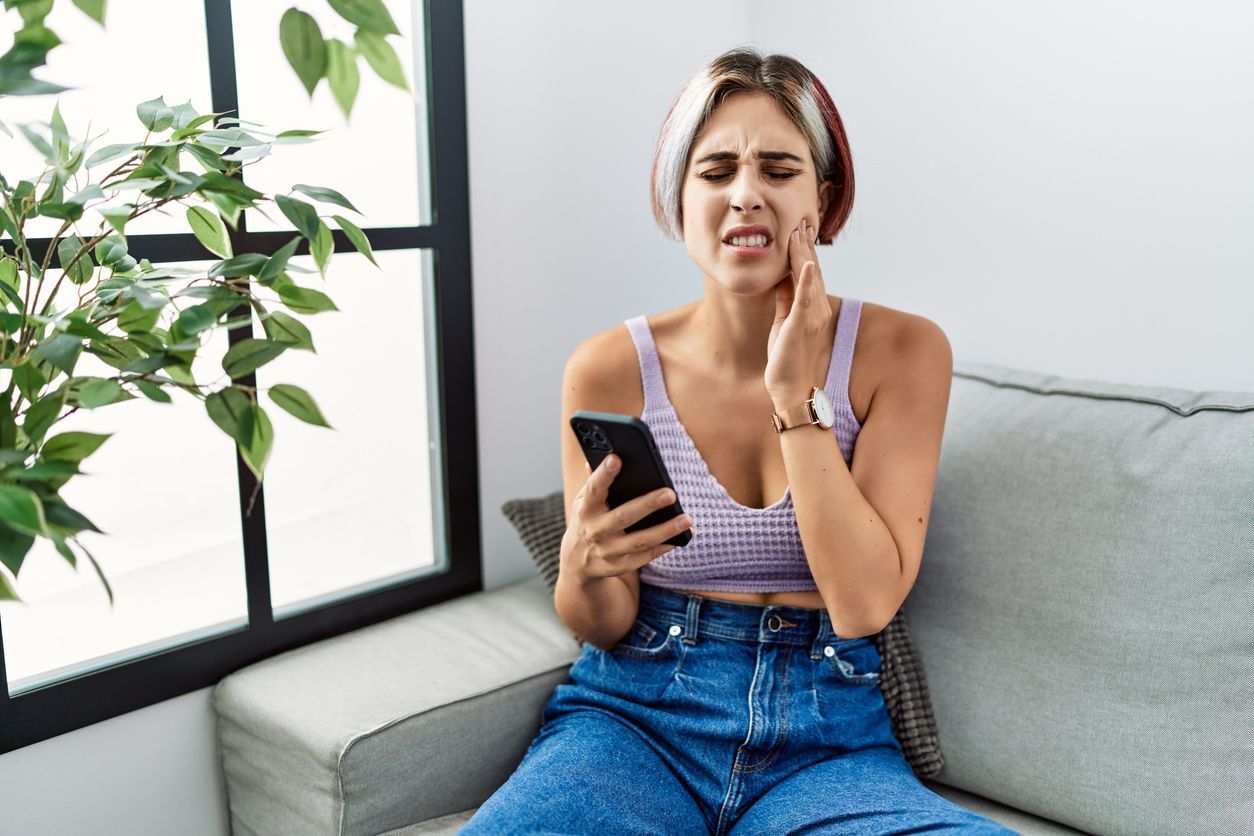 A woman is sitting on a couch holding her tooth while looking at her phone.