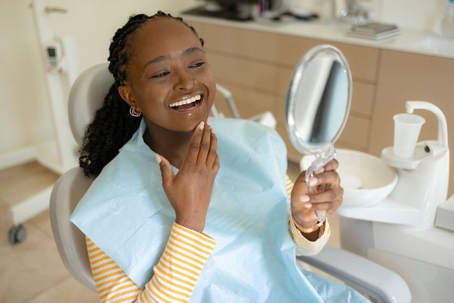 A woman is sitting in a dental chair looking at her teeth in a mirror.