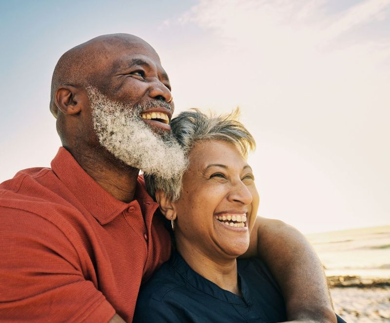 Smiling elderly Black couple embracing at the beach; man with a beard, woman with short hair, bright sunlight.