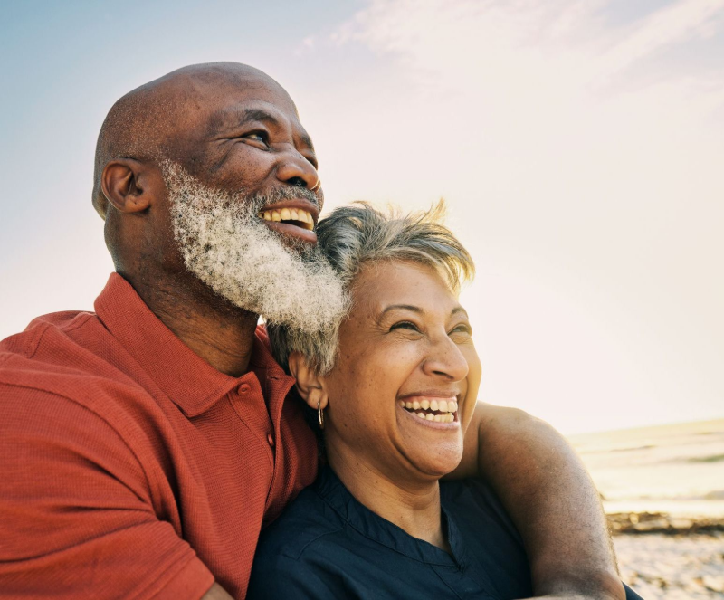Smiling elderly Black couple embracing at the beach; man with a beard, woman with short hair, bright sunlight.