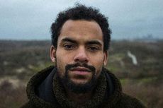Close-up of a man with dark skin and a beard, looking at the camera. He's in a hooded jacket, set against a blurred outdoor background.
