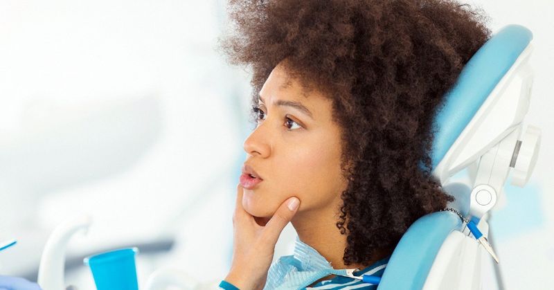 Woman with curly hair in a dentist's chair, touching her jaw with a concerned expression.
