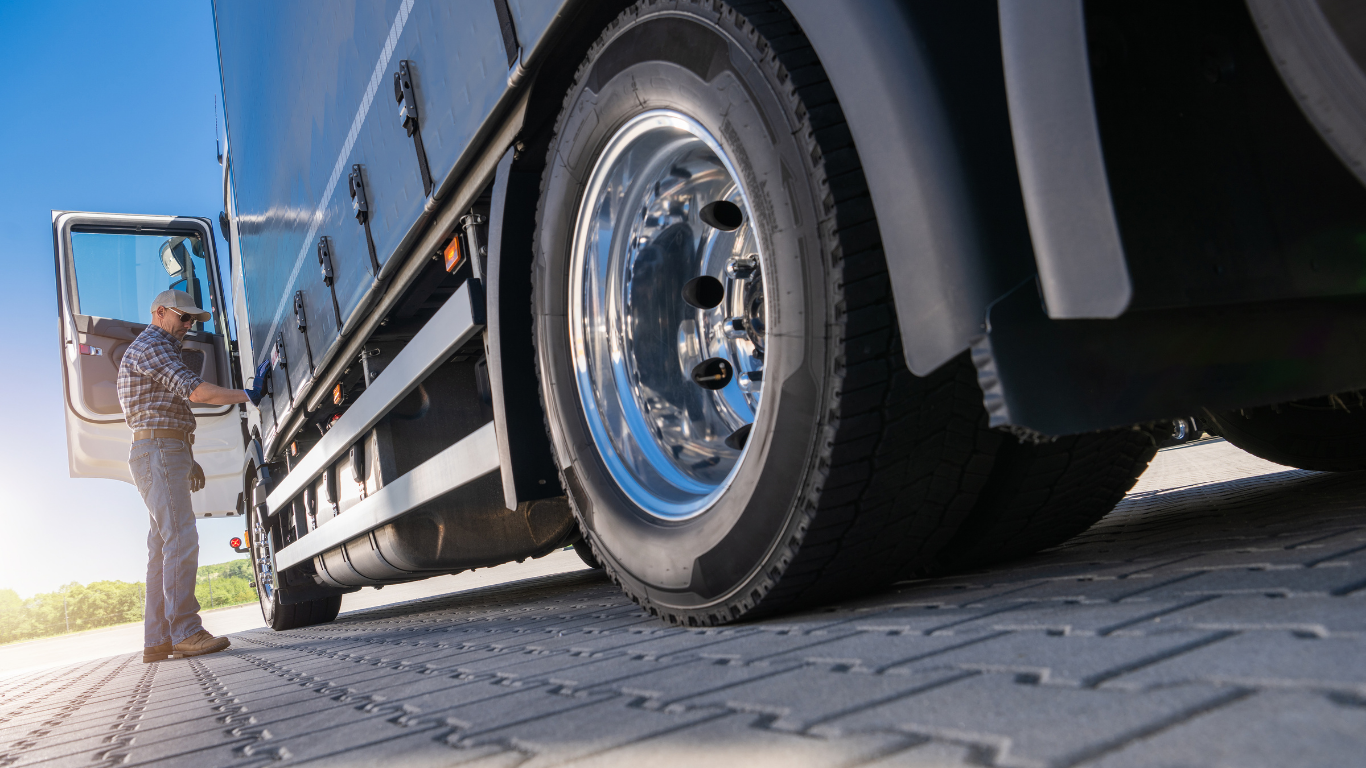 Truck driver standing by a black truck with open door, parked on brick paving, sunny day.