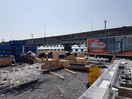 Chantier de construction avec des caisses en bois, des poutres métalliques et un pont en arrière-plan sous un ciel clair.