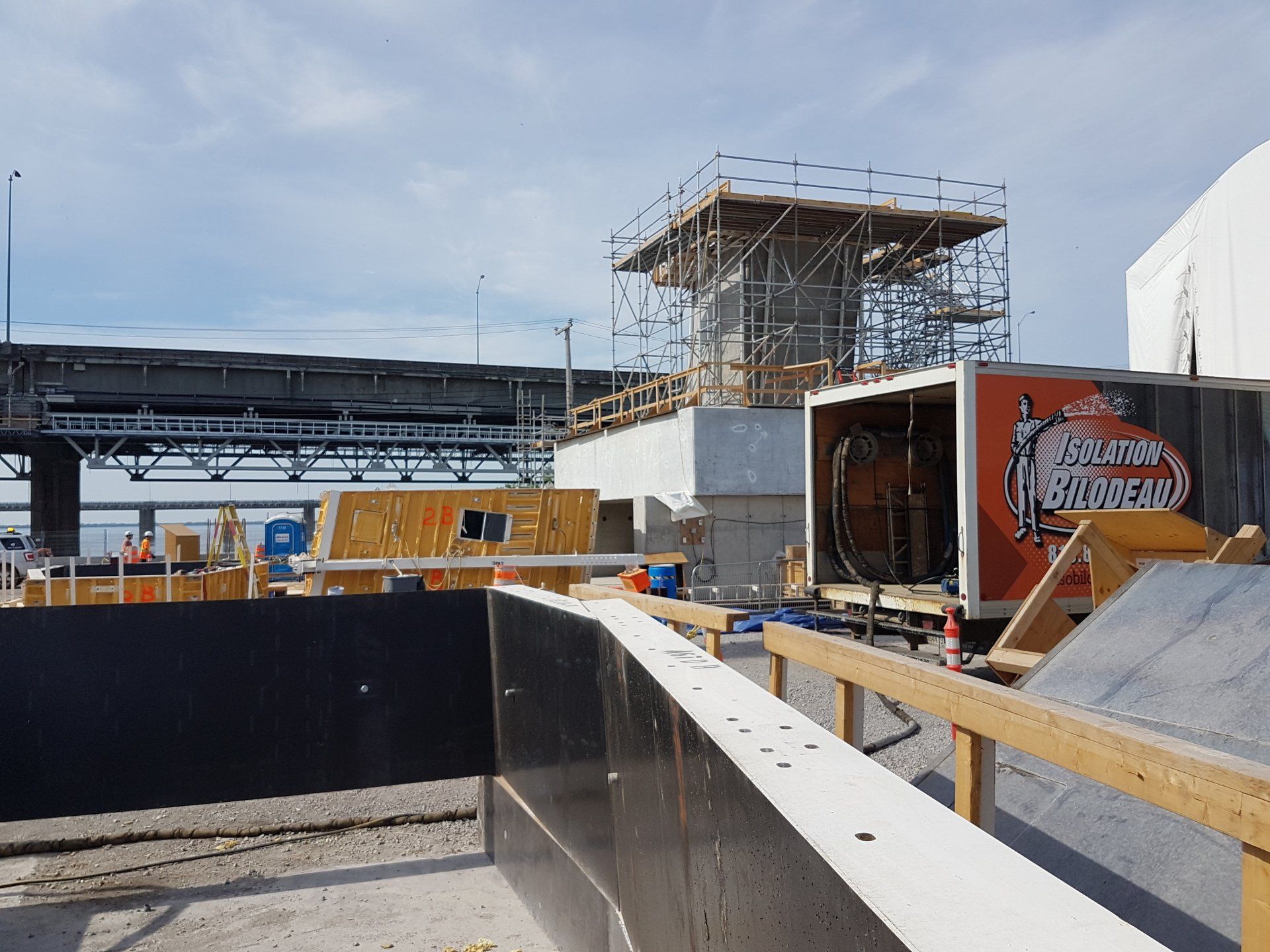 Chantier de construction avec échafaudage entourant une structure en béton, près d'un pont sous un ciel bleu.