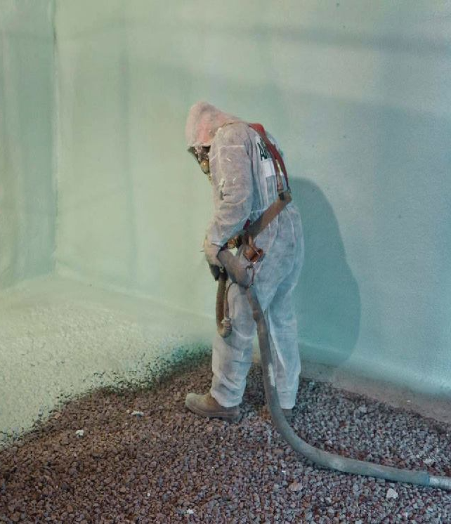 Un ouvrier en combinaison de protection pulvérise du mastic dans une petite pièce aux murs vert clair et au sol en gravier.