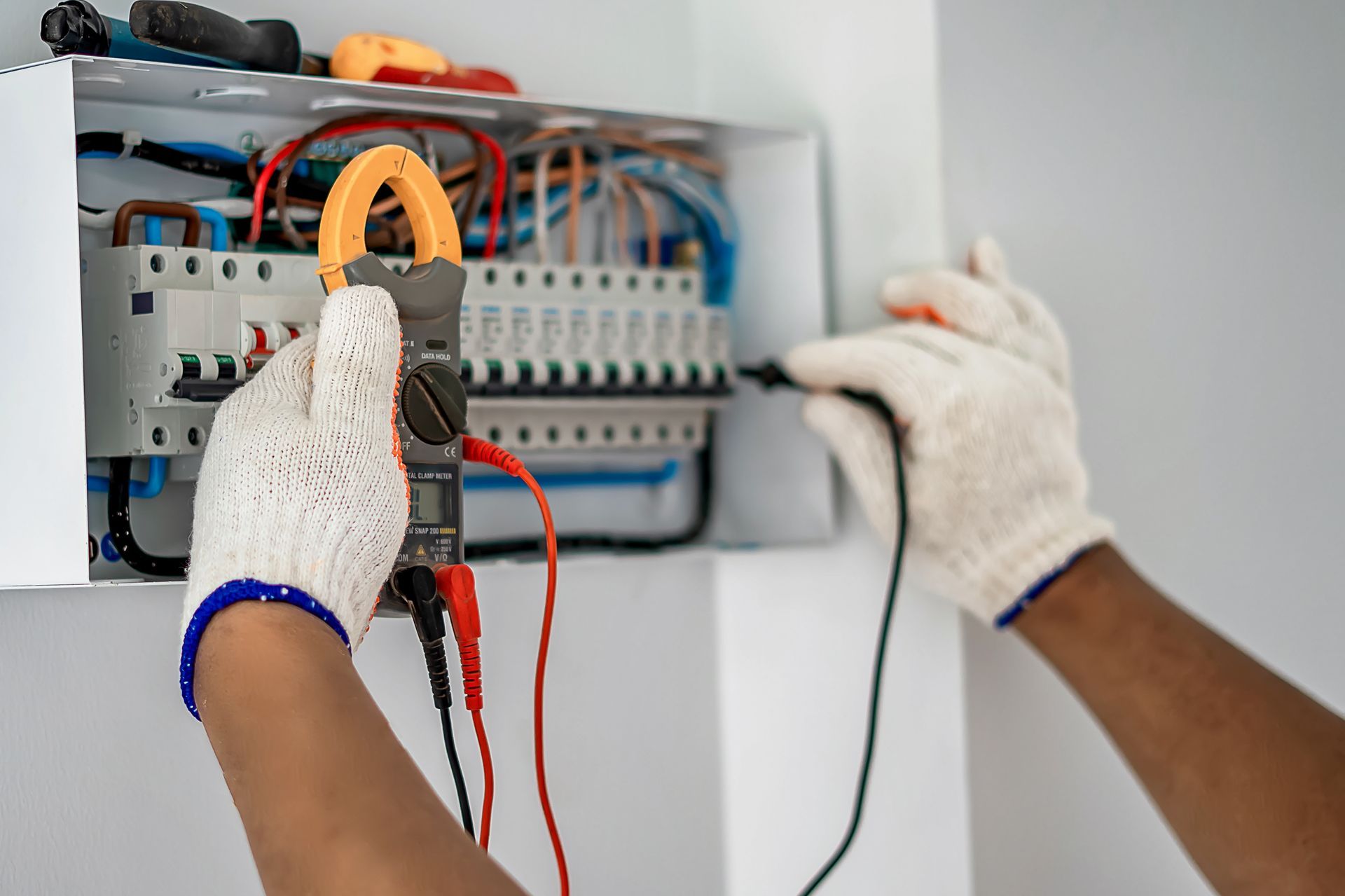 A person wearing work gloves uses a multimeter to test wires inside an open electrical panel.