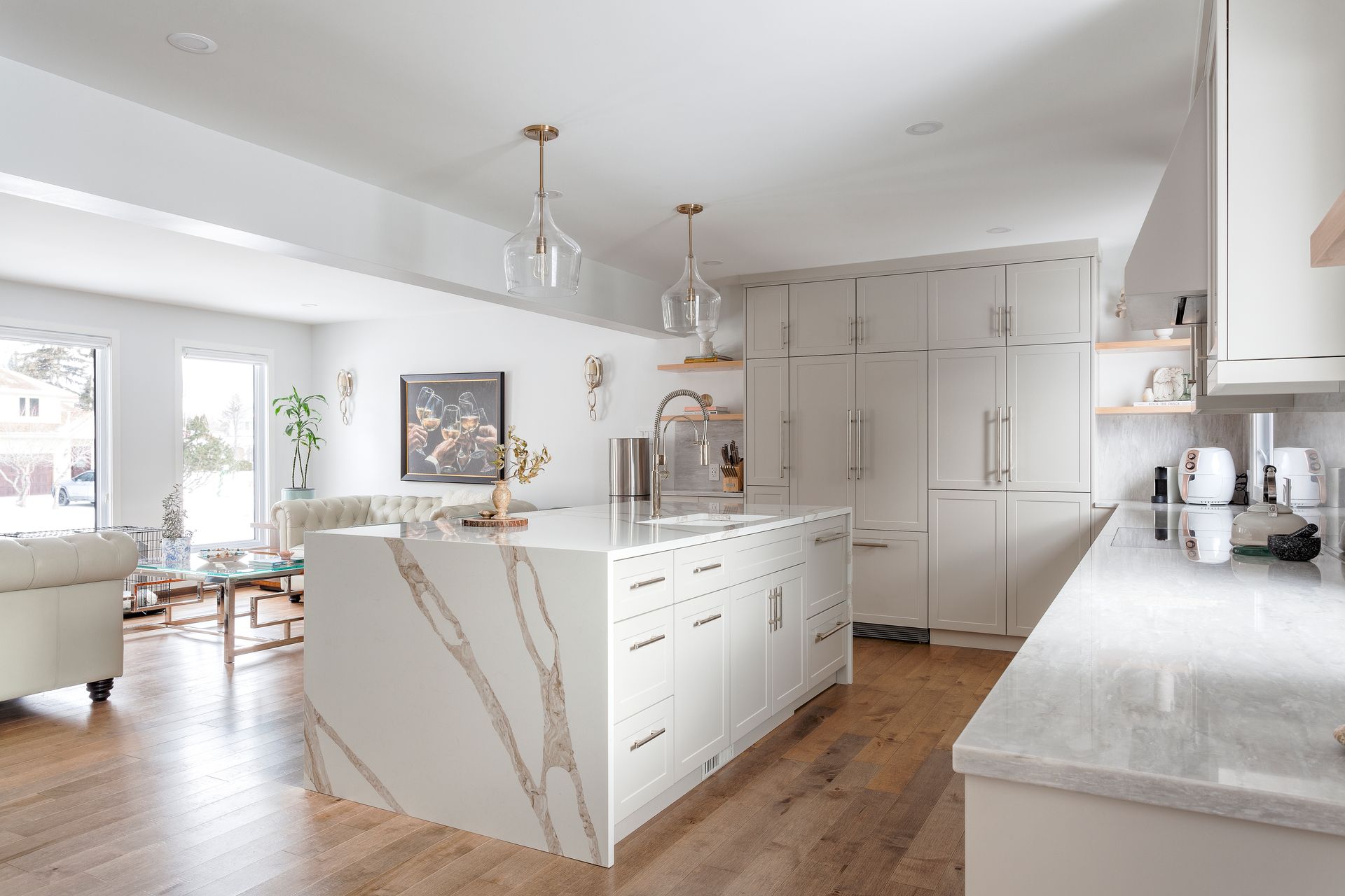Modern white kitchen with quartz countertops and island, connected to living area with hardwood floors.
