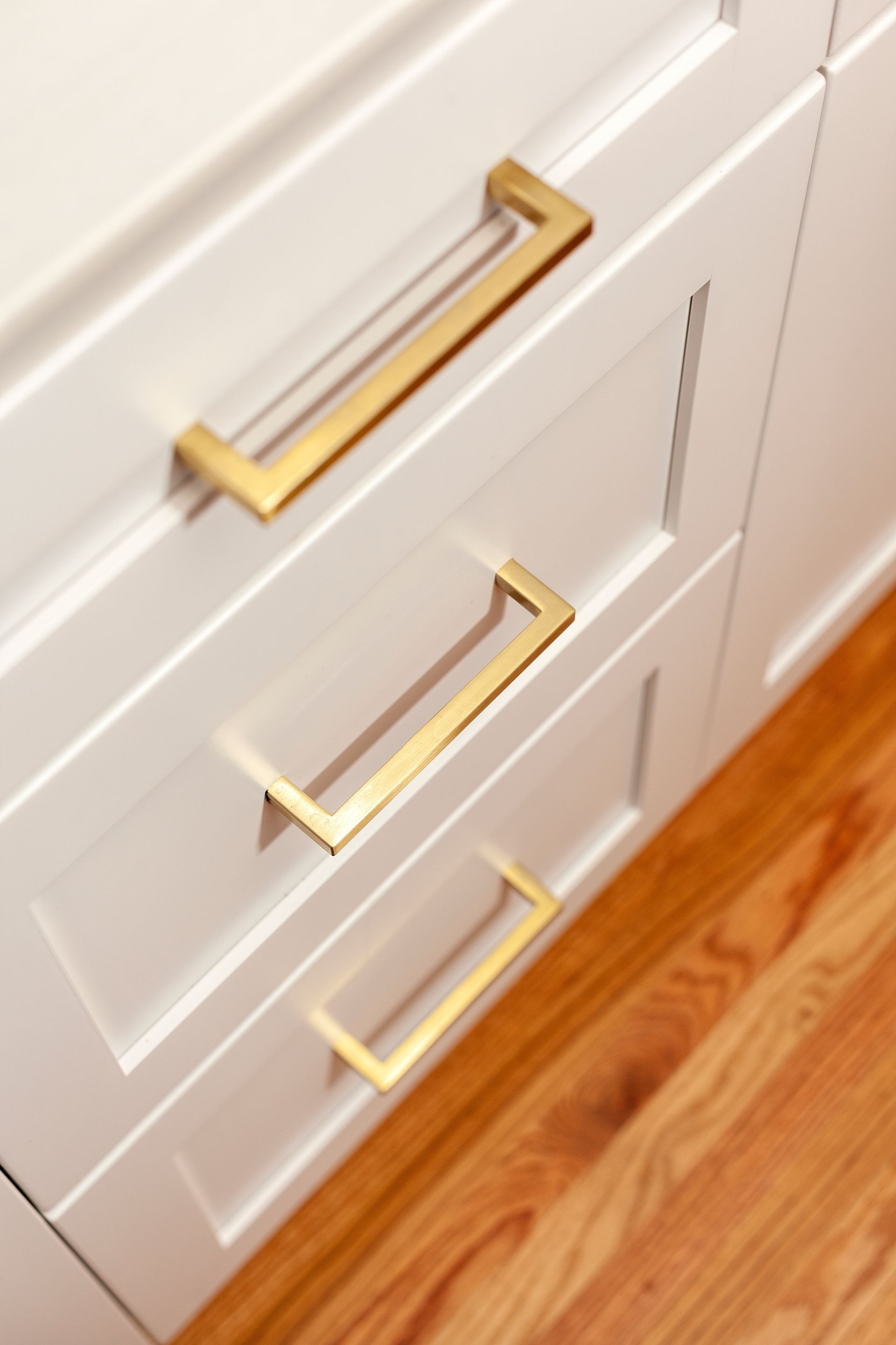 White cabinet drawers with gold rectangular handles, on wooden floor.