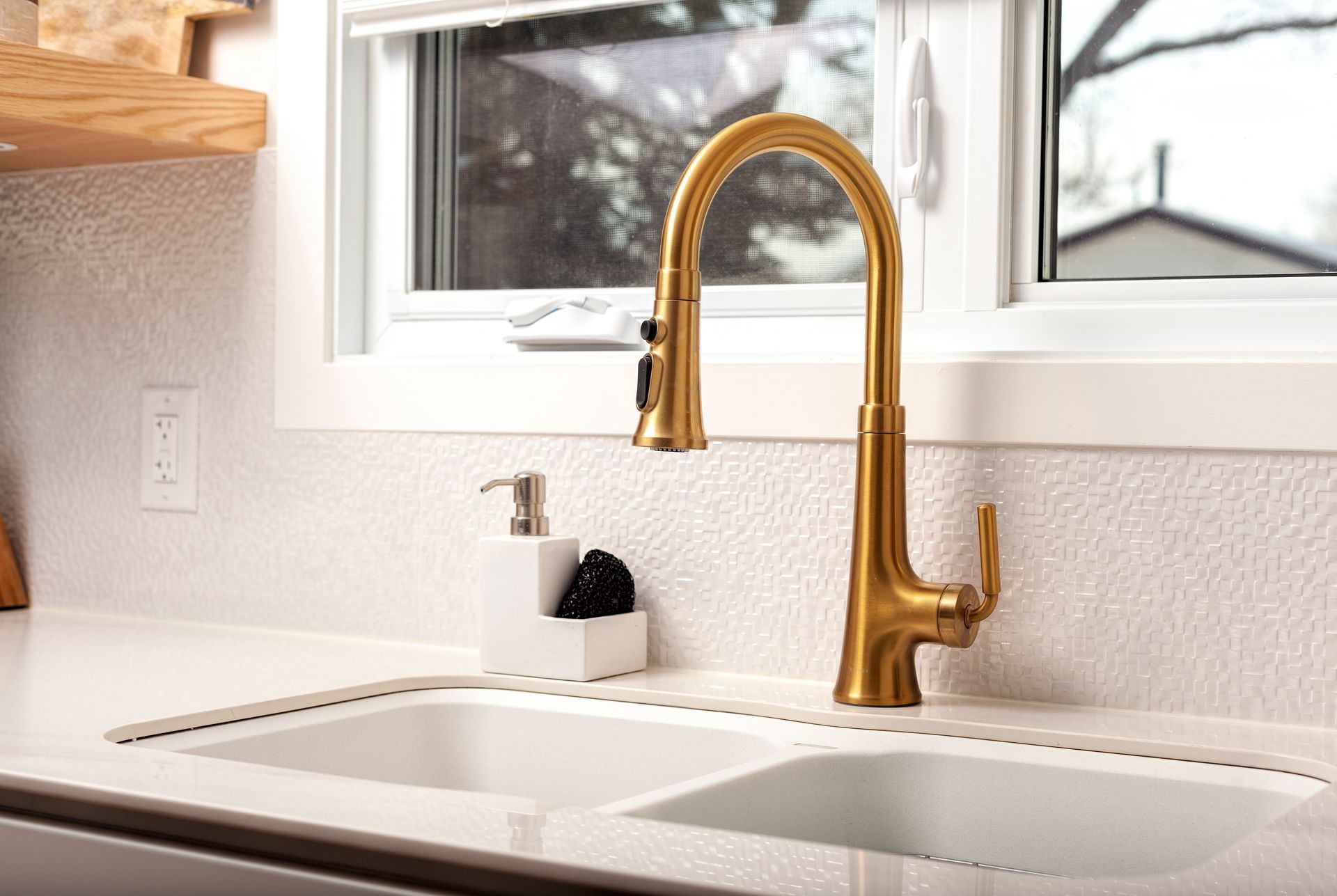 Gold kitchen faucet above a double sink, beside a window. White counters, textured backsplash.