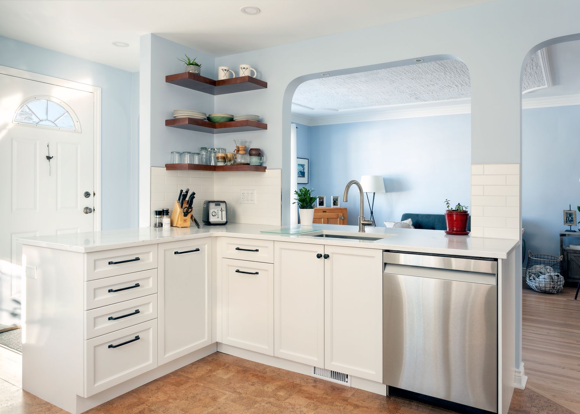 Bright, white kitchen with light blue walls and archway. Includes white cabinets, stainless steel dishwasher, and wooden shelves.