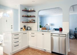 Bright, white kitchen with light blue walls and archway. Includes white cabinets, stainless steel dishwasher, and wooden shelves.