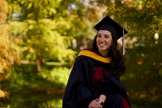 Woman in graduation gown and cap smiling, outdoors near water and trees.