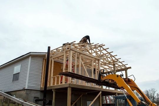 Construction worker on roof of wooden framed addition, using a forklift. Cloudy sky.