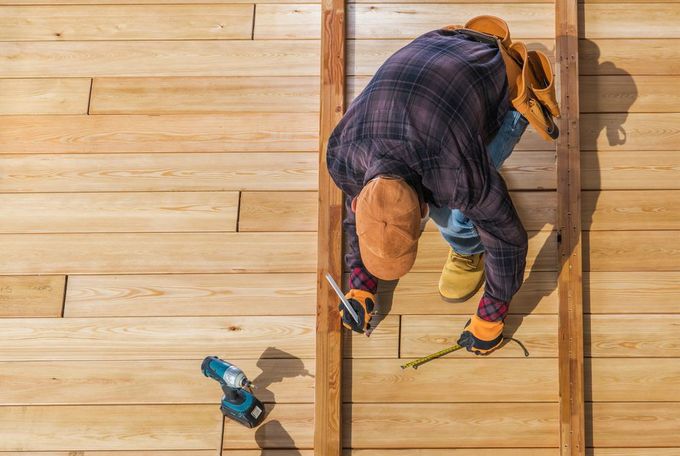 Carpenter measures wood, wearing a plaid shirt, safety glasses, and tool belt. Electric drill on the wood beside him.