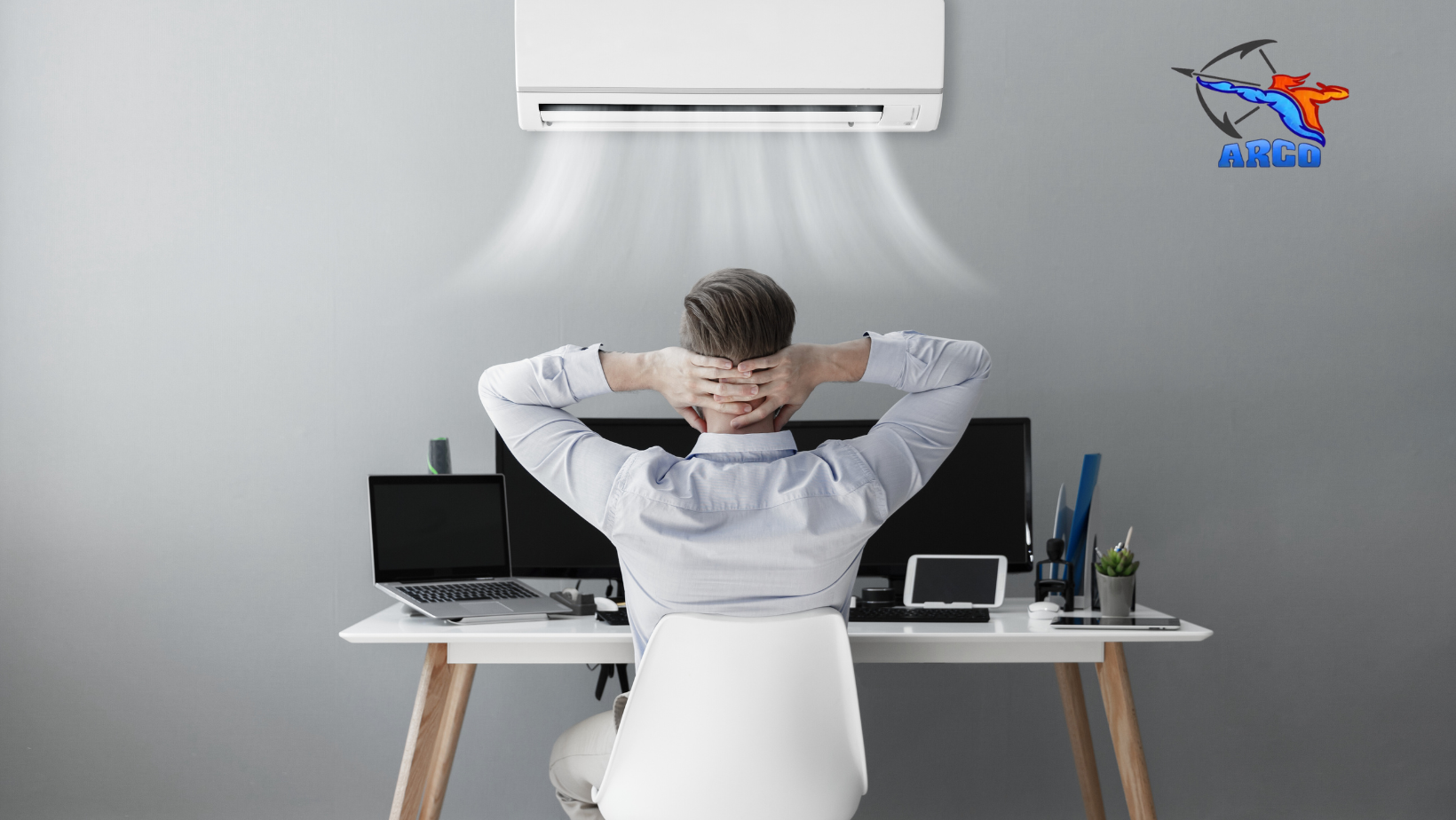 A man is sitting at a desk with his hands behind his head under an air conditioner.