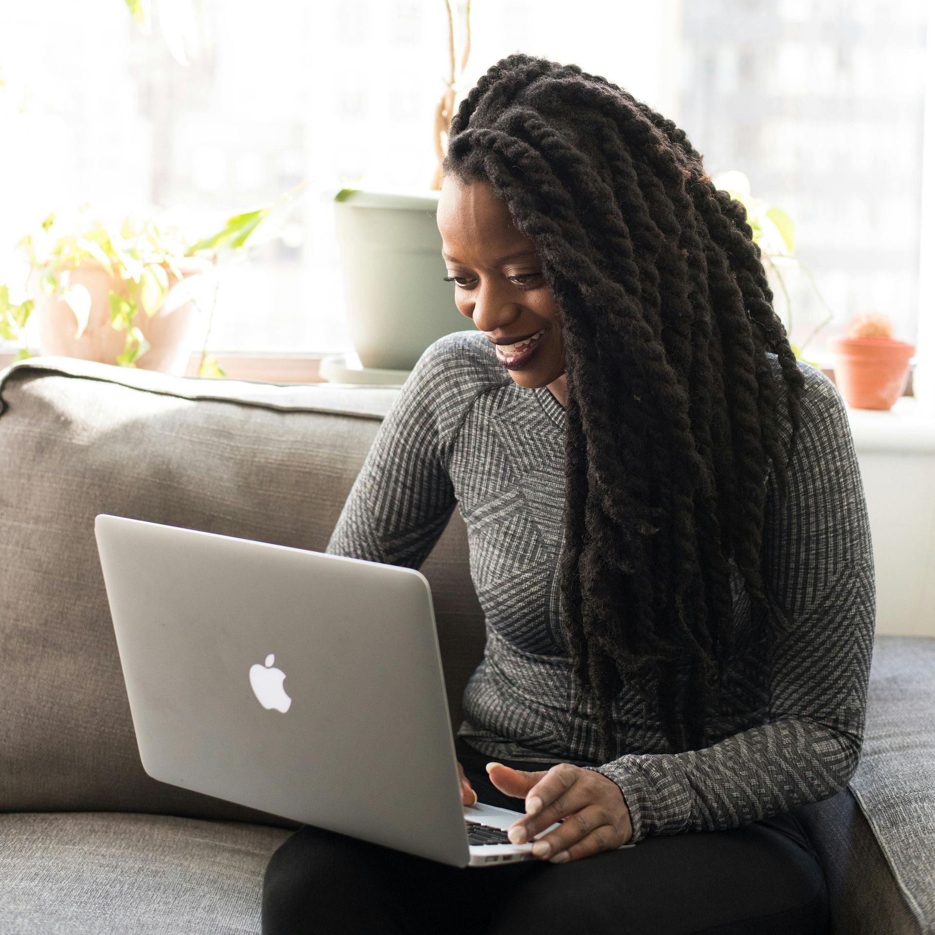 Woman with long braids using a laptop, smiling, sitting on a couch by a window with plants.